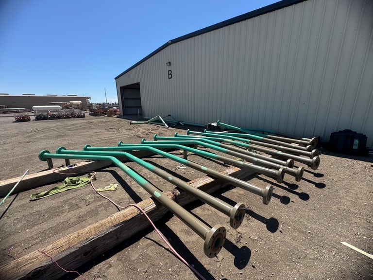 Green and brown irrigation pipe sections stacked in front of a metal industrial building on a dirt lot