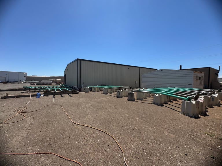 Industrial warehouse buildings with metal siding on a bare dirt lot under clear blue sky, with construction materials and equipment scattered around