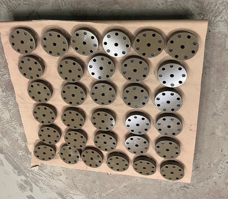 Beige cardboard display board with 36 circular metal discs arranged in rows, most tan with holes, some shiny white
